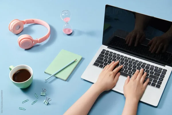 Hands typing on a laptop at a blue desk with headphones, notebook and hourglass